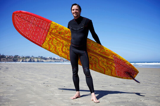 Male surfer holding a colorful surfboard poses for a photo at La Jolla Shores beach.