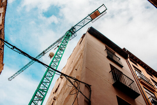 &Aacute;ngulo contrapicado de una gr&uacute;a de construcci&oacute;n verde junto a la fachada de un edificio antiguo bajo un cielo nublado.