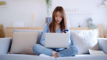 Young asian woman using computer laptop and mobile smartphone while seated on couch at home