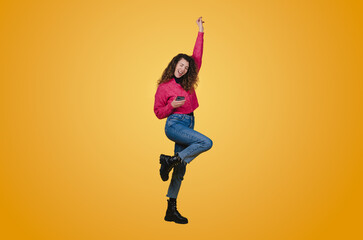 Fototapeta na wymiar Portrait of a happy young woman celebrating victory holding one fist up while holding mobile phone isolated over yellow background. Happy people positive