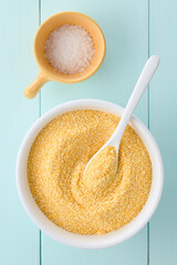 Raw coarsely ground cornmeal or polenta in white bowl, coarse salt on the side, photographed overhead on blue wood (Selective Focus, Focus on the top of the cornmeal)