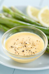 Hollandaise sauce with freshly ground black pepper served in glass bowl, green asparagus and lemon slices in the back (Selective Focus, Focus one third into the image)