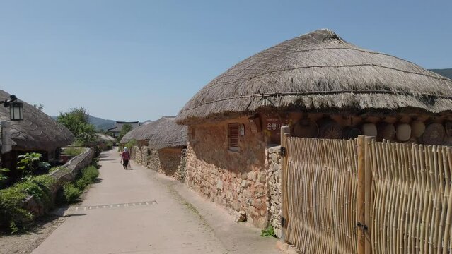 A Village Maintenance Worker With A Broomstick Walking On A Path Between Thatched Roof Houses In Nagan Eupseong Folk Village Near Suncheon Bay, South Korea. - Medium Shot