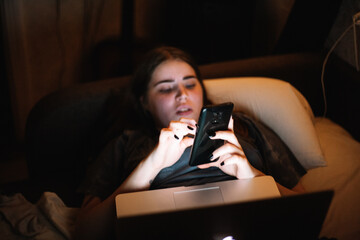 Young woman using smart phone and laptop computer while lying in bed at home at night