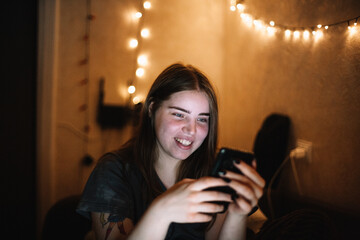 Happy smiling young woman using smart phone sitting in bed at home at night