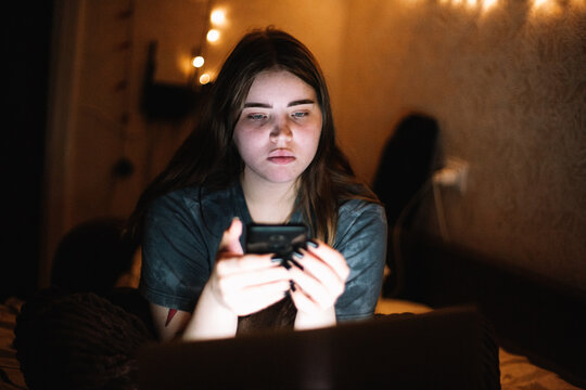 Young female teenage student using smart phone and laptop computer while sitting in bed at home at night