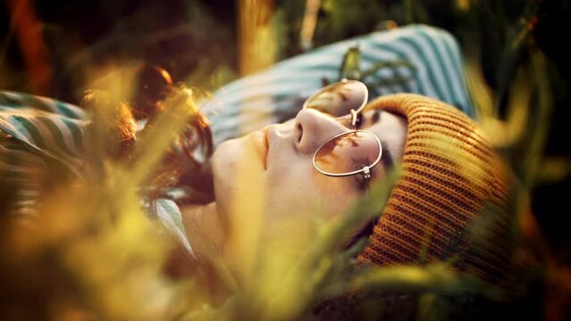 Happy young Carefree hippie woman in sunglasses lying on sunset autumn grass with closed eyes. Pretty girl relaxing outdoor at summertime. Concept of freedom and youth wilderness. Travel vacation.