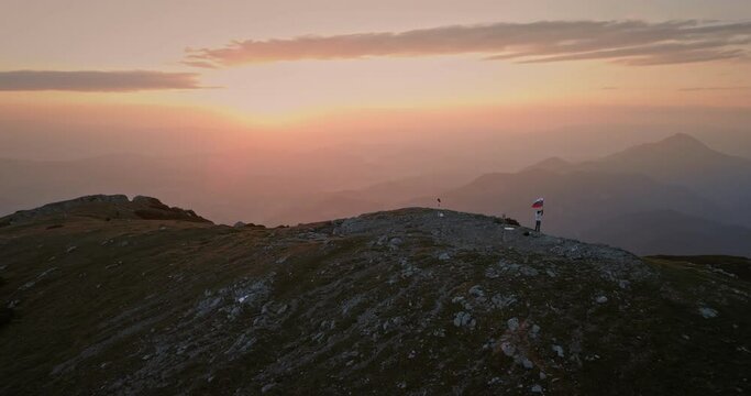 Drone shot of the top of the mountain at the sunrise, hiker holding a slovenian flag on the pol which is fluttering in the breaze.