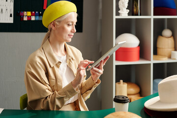 Young creative woman in stylish yellow beret sitting by workplace in design studio and looking for...