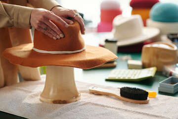 Hands of young craftswoman forming shape of brown felt hat bounded with white decorative shoelace while standing by workplace