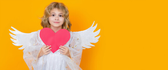 Angel child with heart. Valentine's day. Blonde cute child with angel wings on a yellow studio background. Wide banner panoramic header. Happy angel child.