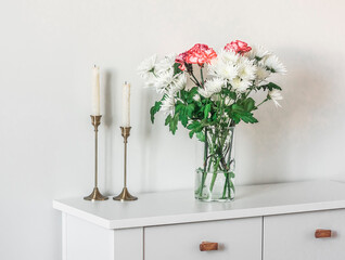 A bouquet of fresh chrysanthemums and carnations, candles in brass candlesticks on a white cabinet in the living room