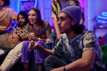 Tense guy in beanie hat and t-shirt looking at tv screen during match broadcast against group of happy multicultural friends