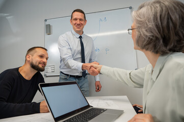 The boss makes a presentation to subordinates at the white board. Caucasian man shaking hands with middle aged woman. 