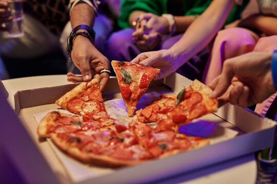 Close-up Of Slices Of Appetizing Pizza In Square Cardboard Box And Hands Of Young Friends Taking Them And Eating While Enjoying Home Party