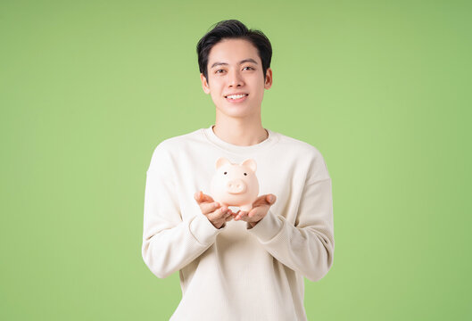 Image of young Asian man holding piggy bank on background