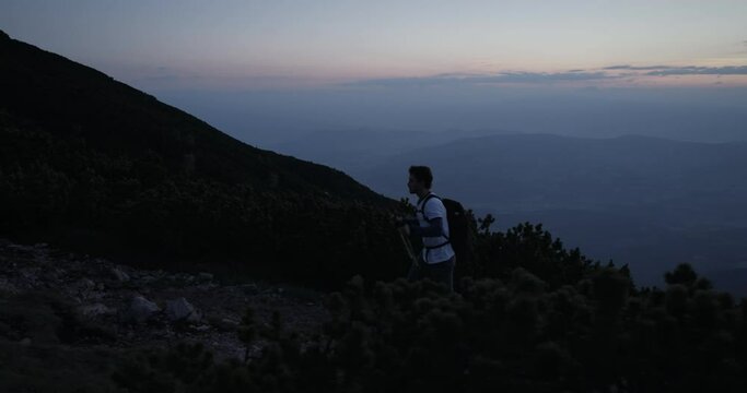 Camera tracking hiker from the side who is walking with hiking polesa up a hill in the dawn on rocky path surrounded with conifers.
