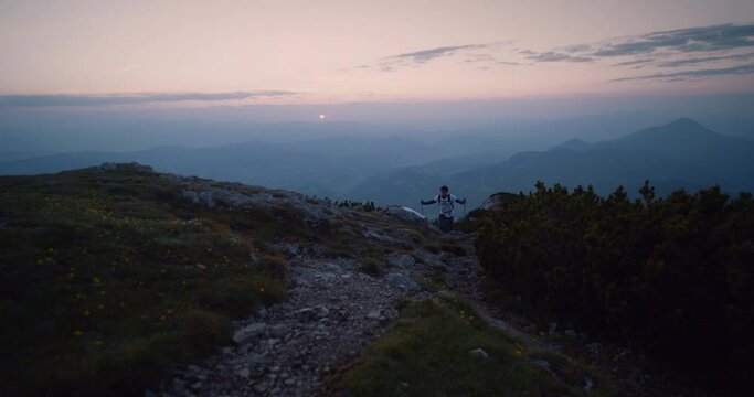 Hiker climbing with the hiking poles up a hill towards the camera in the early morning. A small ball of sunn visible at the horizon.