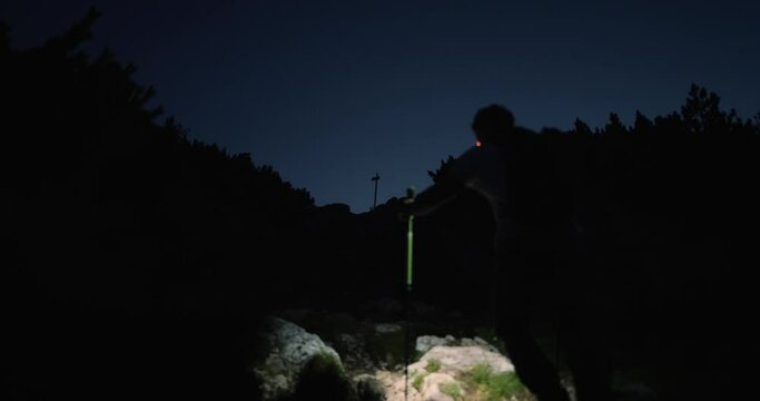 Camera shot from behind of a hiker climbing up a mountain with his headtorch to illuminate his path towards the top. Conifers surronding the path, no clouds on the sky.