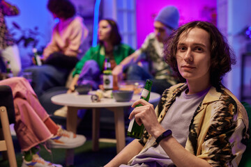 Young man with bottle of beer in hand looking at camera at home party while sitting against his friends chatting and having drinks