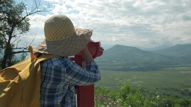 Woman in hat with backpack looking through coin operated binoculars on mountains. Female feels freedom on sunny day. Cheerful woman enjoy outdoor lifestyle on summer vacation trip