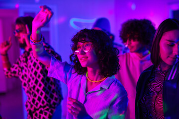 Youthful brunette girl in white blouse and eyeglasses dancing among friends at home party in living room lit by neon light