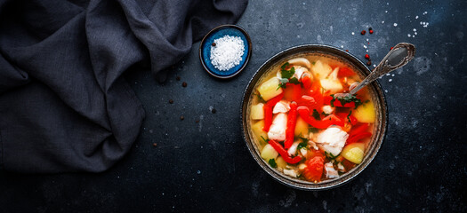 Fish soup with cod, paprika, potatoes, tomatoes and parsley in ceramic soup bowl on dark table background, top view