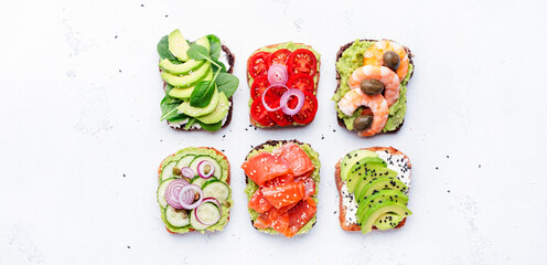 Avocado sandwiches or toast set: with salmon, shrimps, tomatoes, cucumbers, soft cheese and spinach, crushed cashew nuts and sesame seeds, white table background, top view