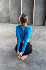 A young woman in sportswear is sitting on a yoga mat with her back and stretching her shoulder muscles. Stretching