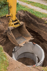 Excavator with a bucket, lowering into the pit on steel cables concrete sewer ring. Construction or...