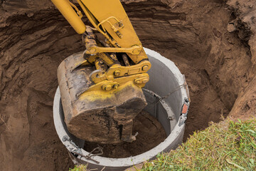 Excavator with a bucket, lowering into the pit on steel cables concrete sewer ring. Construction or...