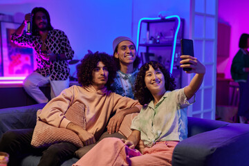 Happy youthful intercultural friends in casualwear taking selfie at home party while relaxing on...