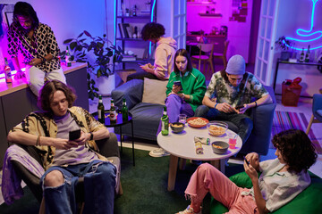 Group of young multicultural friends in casualwear using mobile gadgets while sitting on couch and in armchairs in living room lit by neon light