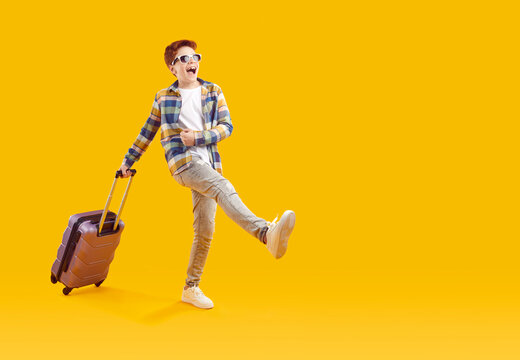 Excited Happy Teenage Boy In Sunglasses Walking With Suitcase. Full Length Shot Of Tourist Boy Wearing In Plaid Shirt And Jeans Traveling With Luggage On Vacation On Isolated Yellow Studio Background