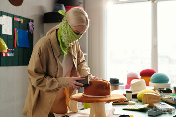 Young creative woman with bandana on face polishing new handmade felt hat on special equipment...