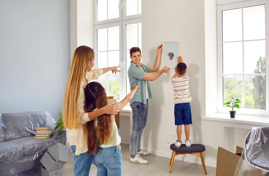 Family is settling in new house. Joyful man and his little son together hang picture on wall under guidance of woman and daughter. Young family hangs picture together while moving to new apartment.