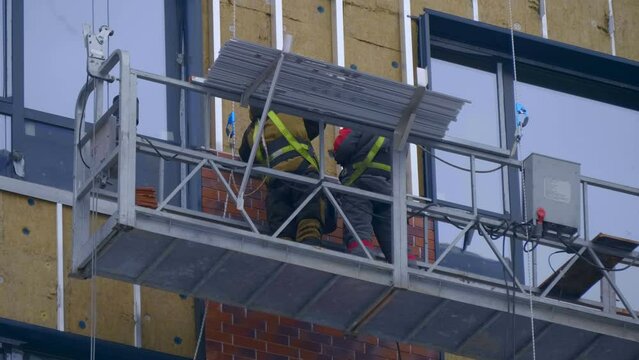 At a construction site, builders in a cradle on cables fastens decorative cladding panels on the wall of a building under construction