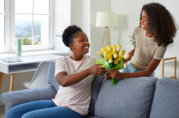 Happy, smiling child gives flowers to mom on Mother's Day. Little African American daughter gives bouquet of beautiful yellow tulips to her mommy who is sitting on sofa in living room