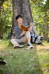 Handsome Asian male college student reading a book under the tree in greenery park.