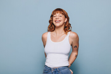 Cheerful young woman smiling at the camera while standing in a tank top and jeans - obrazy, fototapety, plakaty