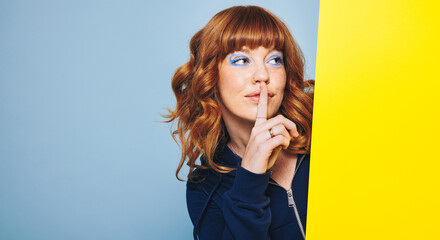 Woman with ginger hair and blue eyeshadow making a hush gesture behind a placard