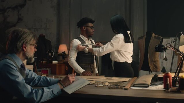 Female tailor measures African American client, discusses with male tailor design of tailored suit. Man sits at table with tailoring tools and making notes. Concept of fashion and hand craft.