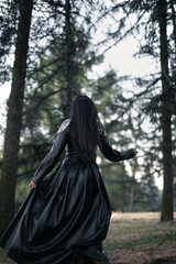Naklejka premium Dark victorian aesthetic scenic photography. Picture of woman goth in black dress with spruce forest on background. High quality wallpaper. Photo concept for ads, travel blog, magazine, article