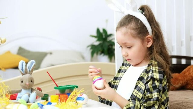 Little girl dyeing Easter eggs. Child painting egg for Easter hunt. Home decoration with flowers, basket and rabbit for spring holiday celebration.