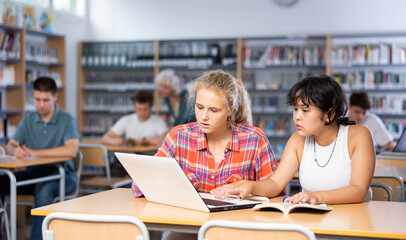 Portrait of asian schoolgirl with her spanish female friend studying in library using laptop