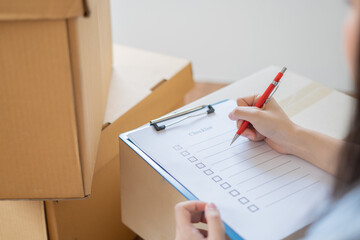 Asian woman checking and packing the carton box prepare to move to new house relocation shipping or check goods package before delivery to customer from online shopping by checklist
