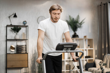 Fototapeta na wymiar Caucasian muscular sporty man in white t-shirt leaned his hands on treadmill while working out in morning at living room at home.
