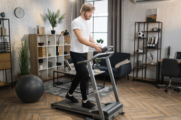 Fototapeta premium Full length view of blond sporty man in white t-shirt and black sports pants having cardio training to loss weight using treadmill at home gym.