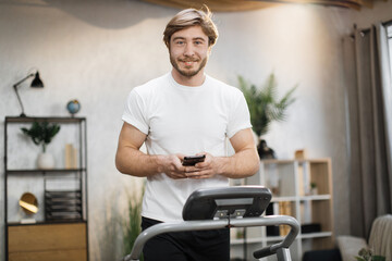 Handsome young sports man using phone looking online fitness class while working out, running, doing cardio training on treadmill in morning time, indoor on background of modern apartment