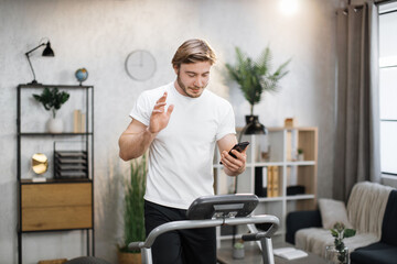 Front view of caucasian man in sports clothes using smart phone for video call doing cardio training on treadmill at home or gym. Concept of sport, health care, action, remote leisure.